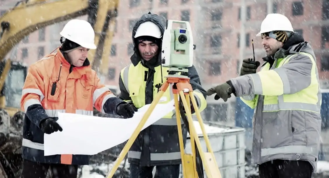 Three construciton workers around plans and a laser level in the snow on a job site
