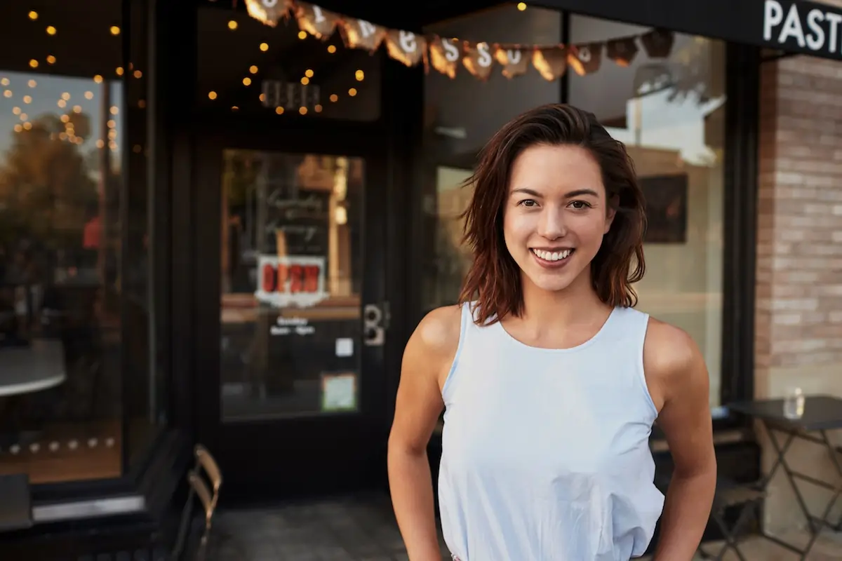 Woman shop owner standing on sidewalk in front of shop looking into camera and smiling